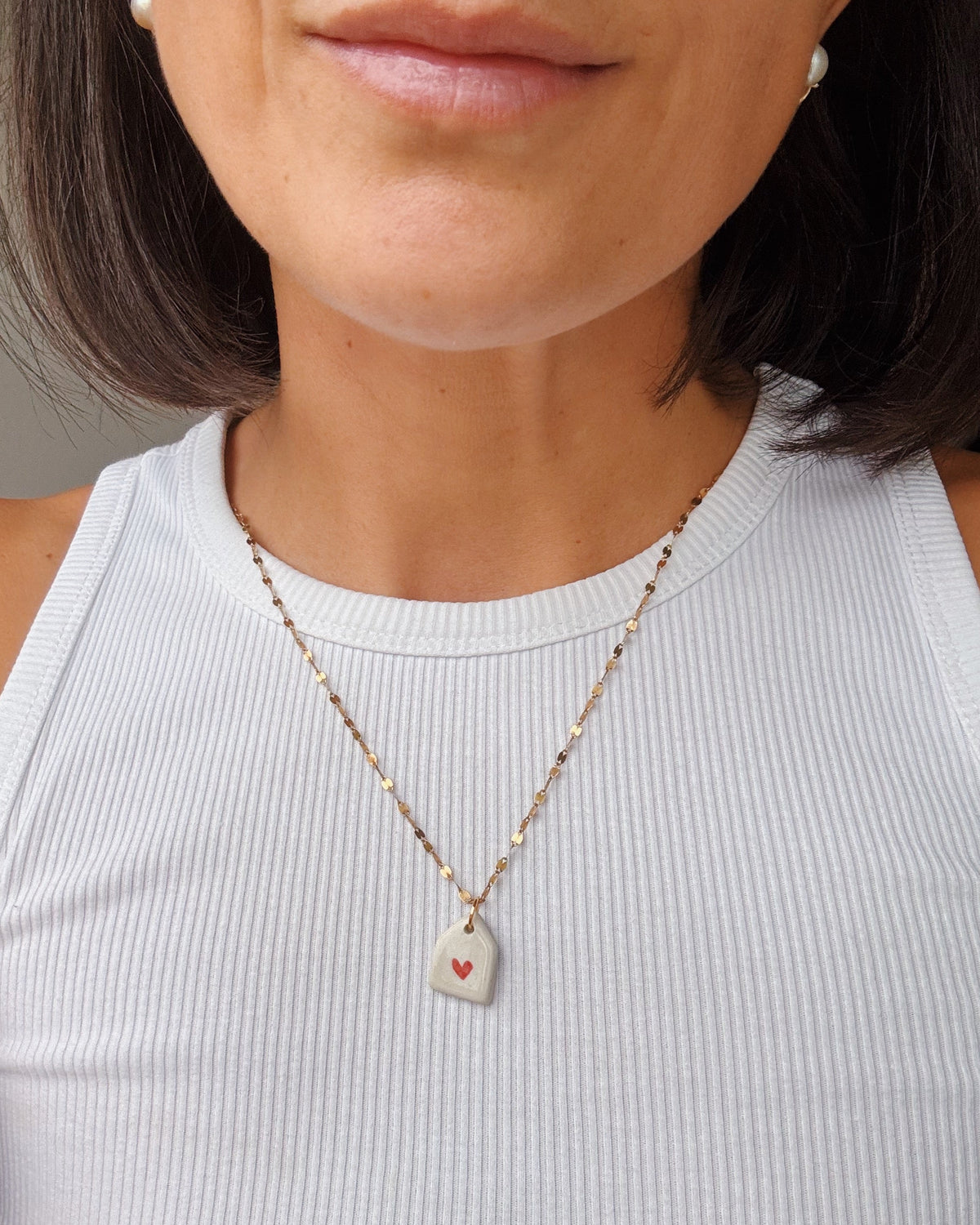 A close-up of a woman wearing a delicate gold chain necklace with a small white guitar-pick-shaped pendant featuring a tiny red heart, paired with a white ribbed tank top.