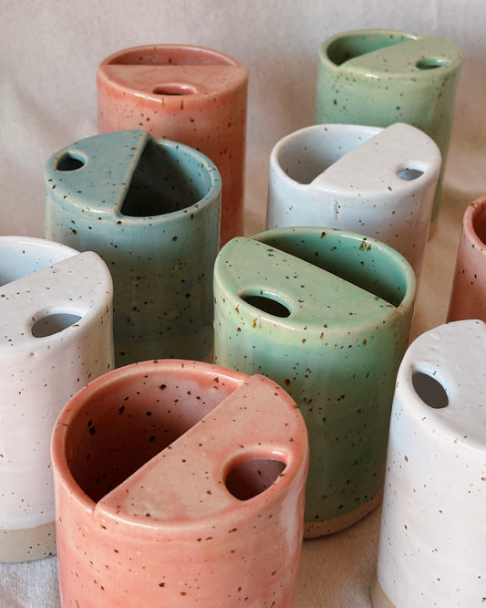 A close-up of several ceramic travel mugs shown in multiple pastel colors including white, mint green, light blue and pink. The cups have a speckled glaze and are arranged overlapping on a light fabric background.