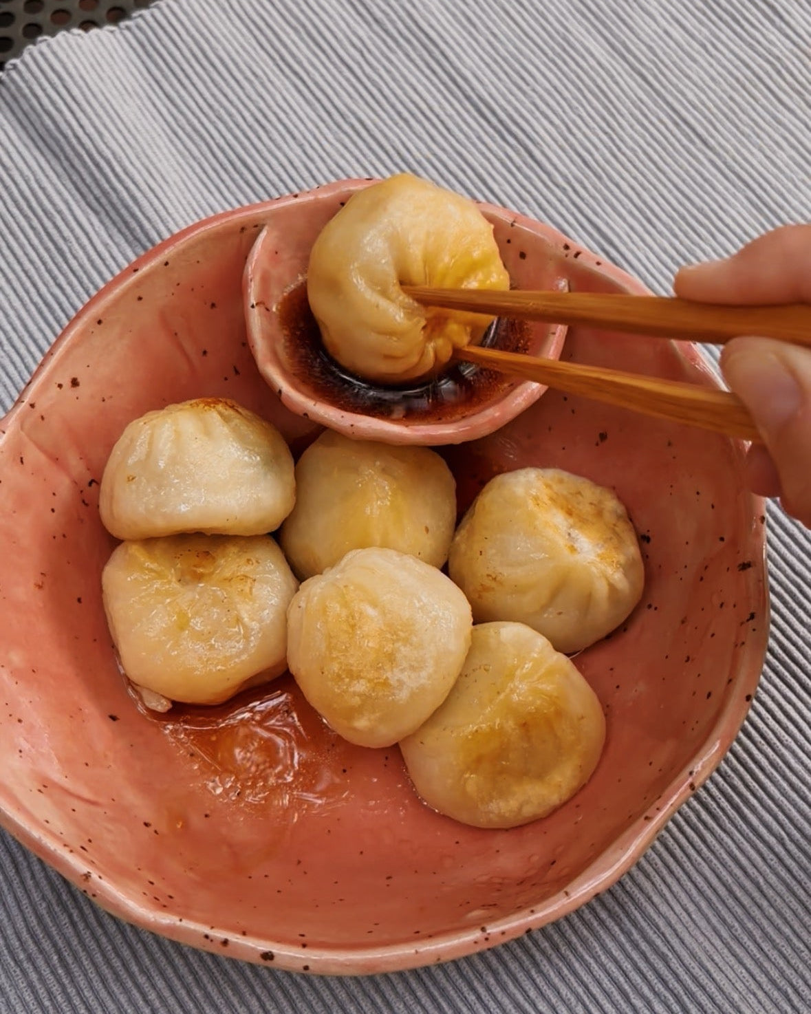 A close-up of golden-brown pan-fried dumplings in a speckled terracotta bowl, with chopsticks lifting one dumpling over a dipping sauce, set on a striped cloth.