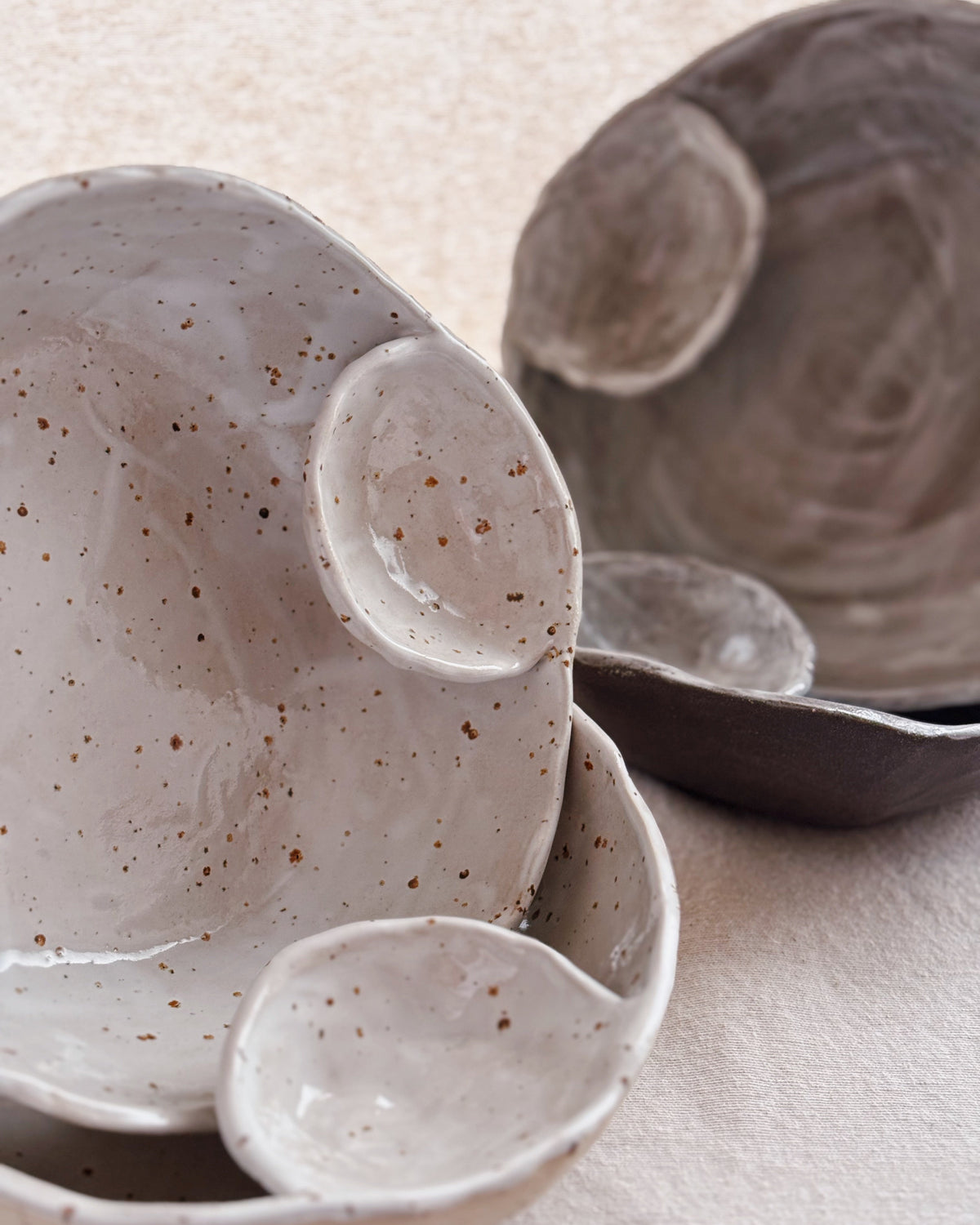 Handmade ceramic bowls with a speckled glaze in light gray and dark gray tones, arranged on a textured beige fabric background.