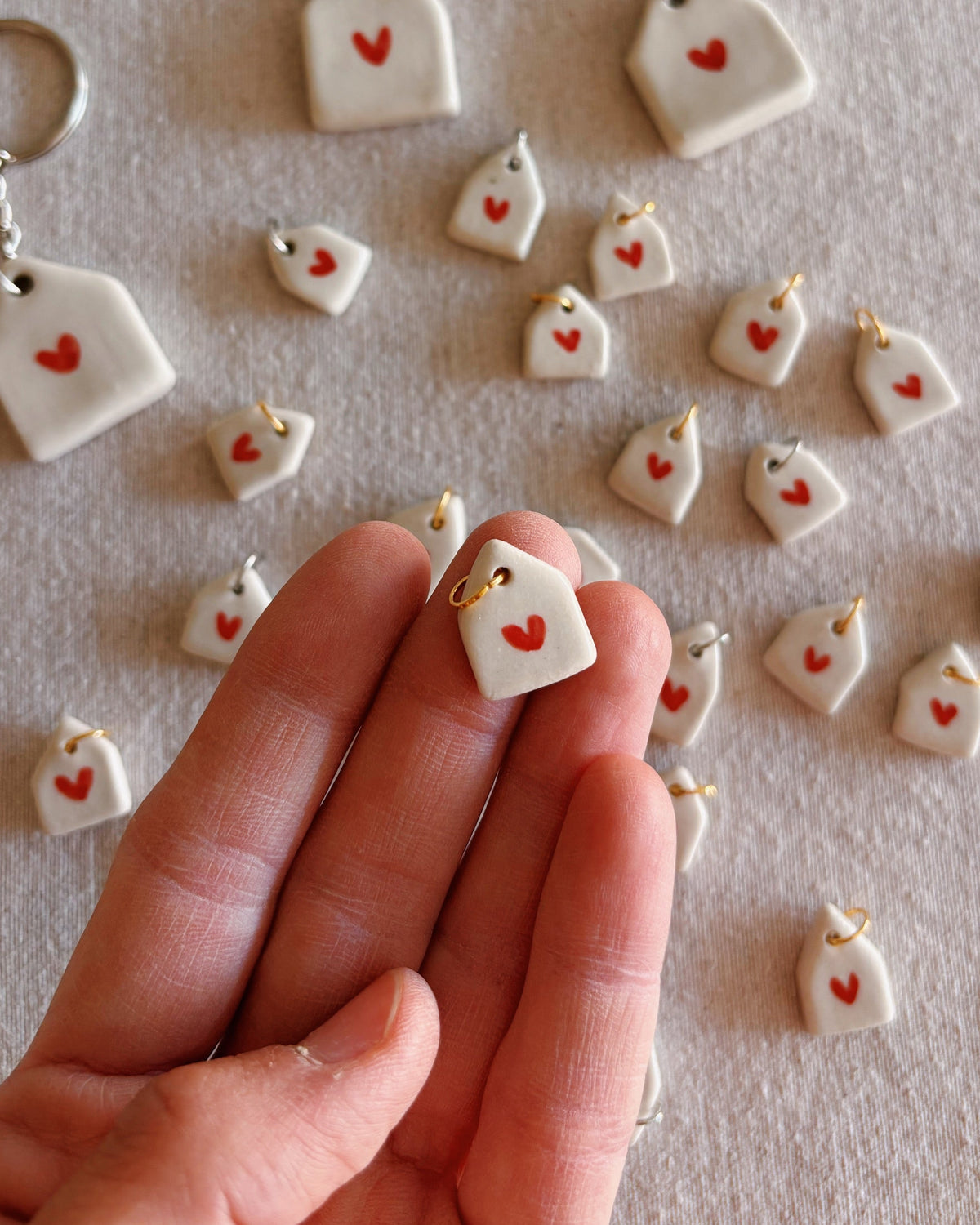 A hand holds a small white ceramic keychain shaped like a house with a red heart, surrounded by many matching keychains scattered on a light beige textured surface.