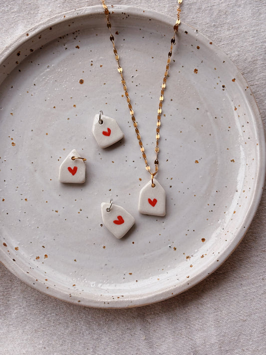 A gold chain necklace and matching earrings with small white ceramic pendants decorated with tiny red hearts, displayed on a speckled ceramic plate over a neutral fabric background.