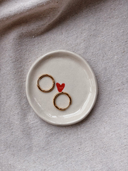 Two thin gold rings displayed on a small round off-white ceramic dish with a red heart in the center, placed on a textured beige fabric background.