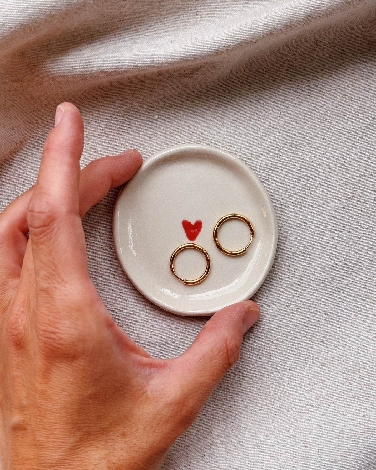 A hand holds a small round white dish with two gold rings and a tiny red heart on a textured beige fabric background.