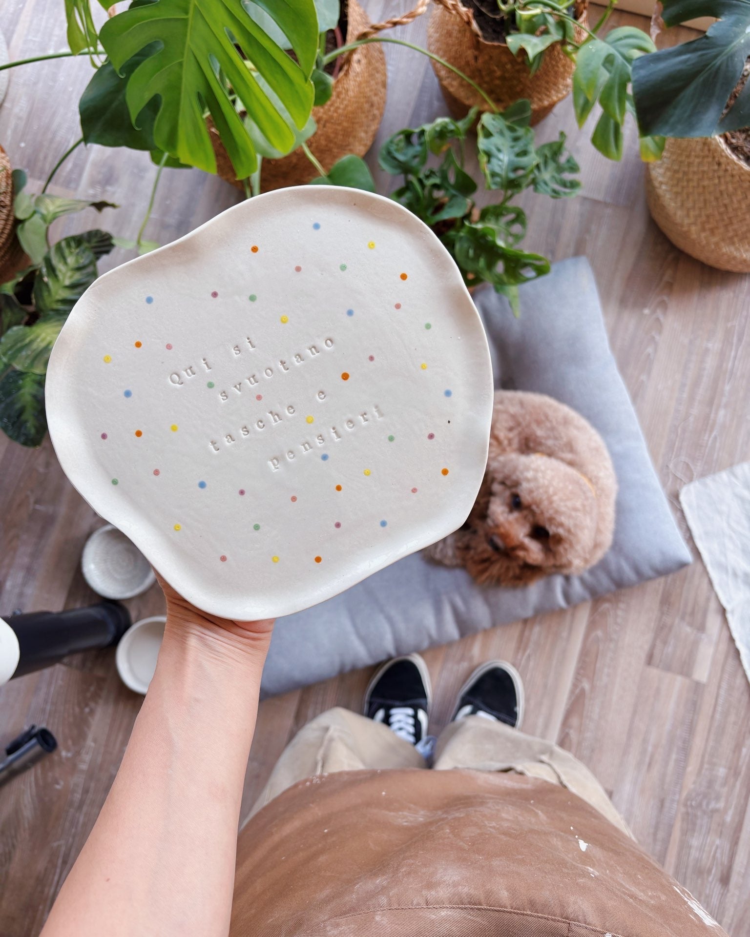 Person holding a decorative plate with text and colorful dots in a home setting with plants and a dog.