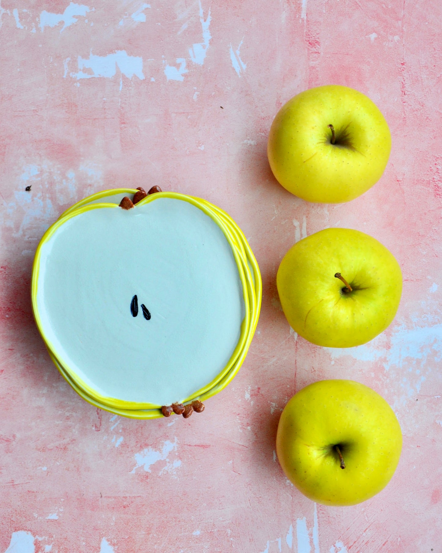 Apple Spoon Rest in the shape of an apple, surrounded by fresh yellow apples on a pink background.
