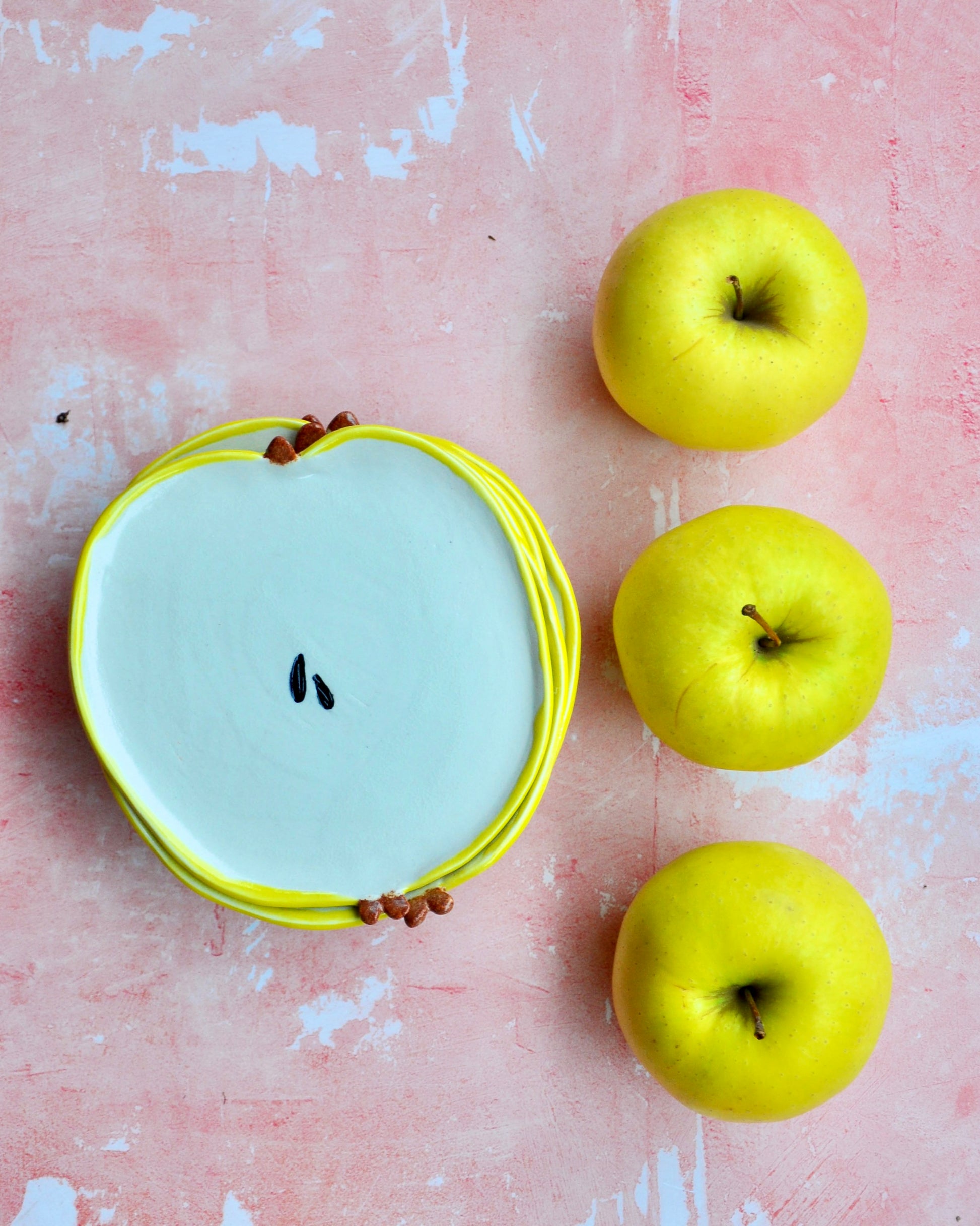 Apple Spoon Rest in the shape of an apple, surrounded by fresh yellow apples on a pink background.