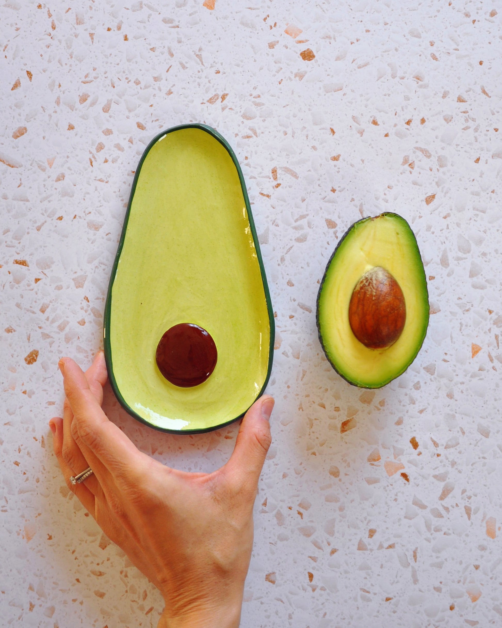 Avocado Spoon Rest handmade stoneware, showing a hand holding the spoon rest next to a half avocado.